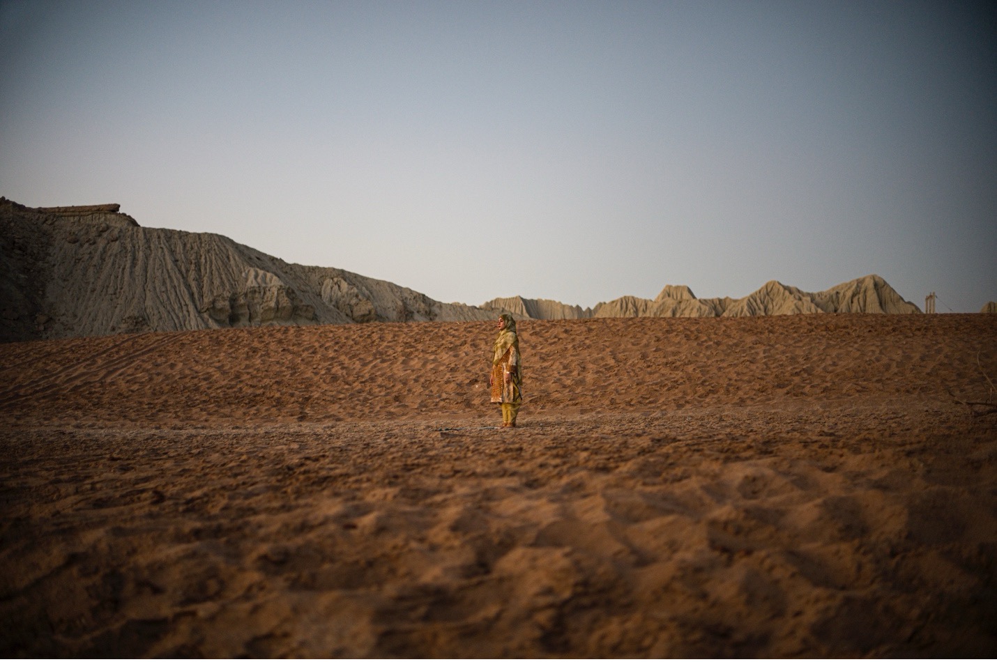 A side profile of a woman standing in the distance. The woman is wearing a tan dress and looking off to the left. She is standing in an arid landscape. The ground is sand, and no vegetation is visible. In the distance, hills are visible. The sky is clear and fades to black in the corners.
