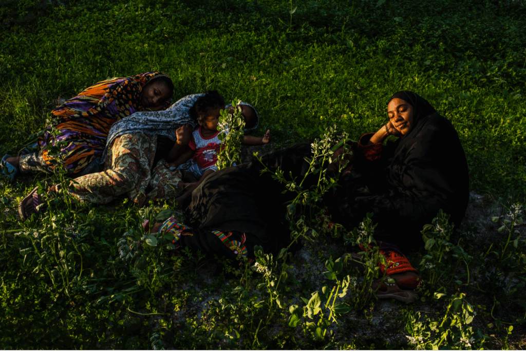 Three brown-skinned women and a child lounge in leafy vegetation. the woman closest to the camera has her eyes closed and her head resting on her hand.