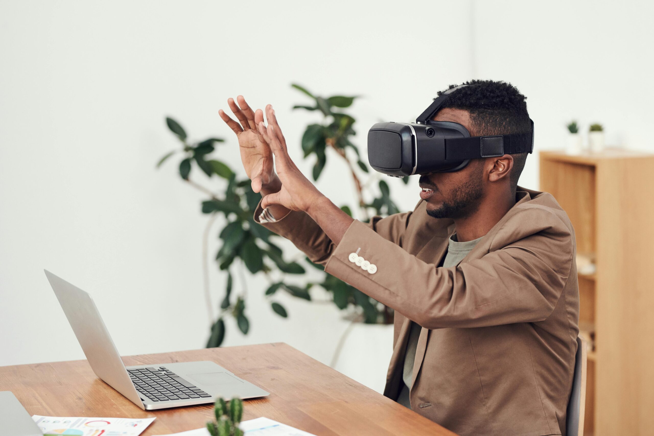 A Black man wears a light brown suit and virtual reality goggles. He is seated in front of a laptop with a bookcase and plant visible behind him. He is holding his hands in front of his face.