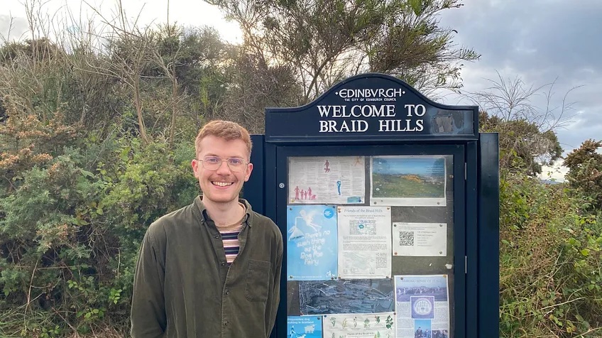 Cllr Ben Parker standing in front of the sign and noticeboard at the entrance to Braid Hills park.