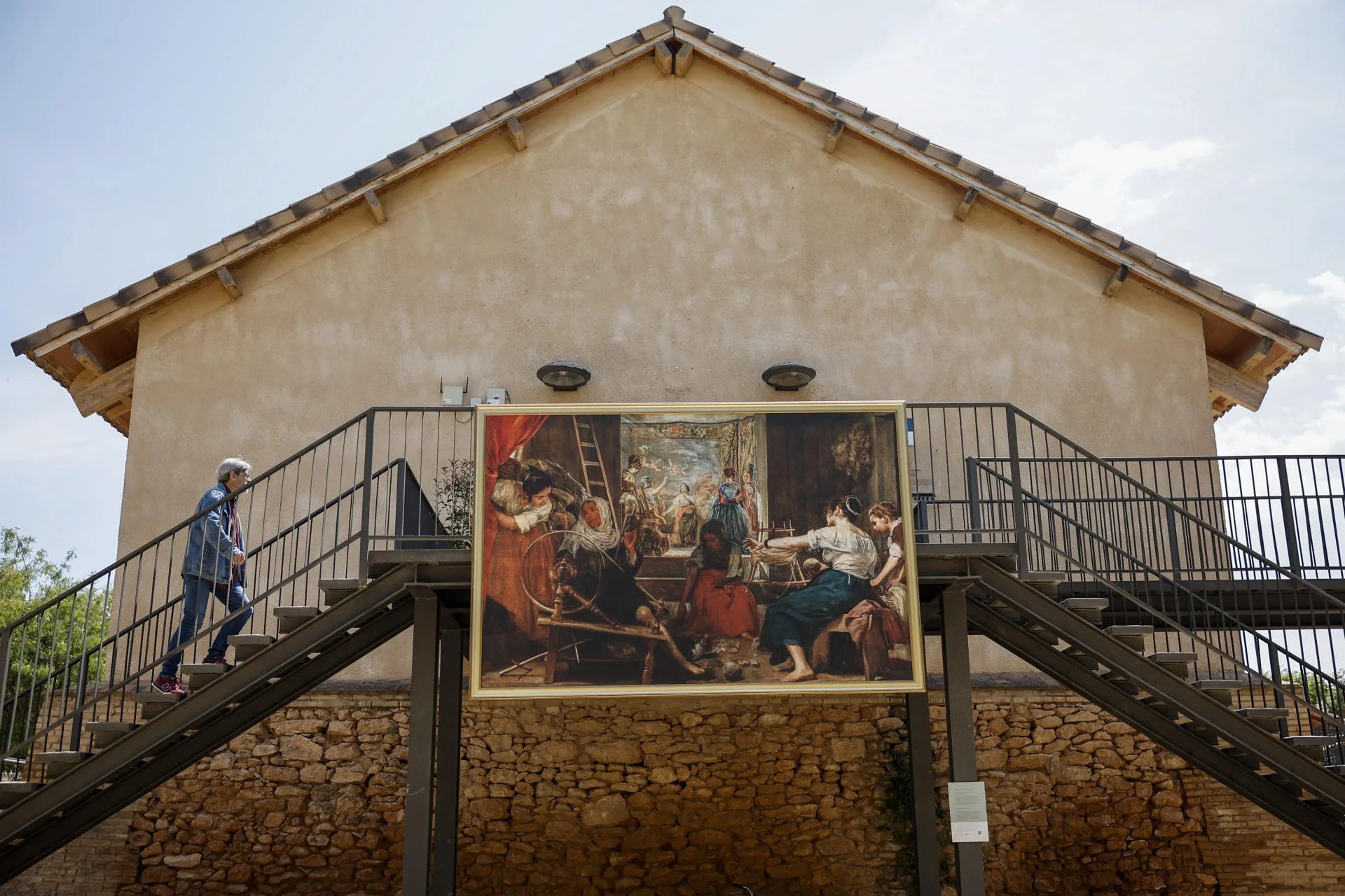 GRAFCVA7472. PAIPORTA (VALENCIA), 13/05/2025.- Una persona pasa ante el cuadro de Diego Velázquez "Las hilanderas" o "La fábula de Aracne" durante la presentación de la iniciativa del Museo Nacional del Prado En un lugar de… , que convierte calles y plazas de 12 localidades de Valencia y 1 municipio de Castilla la Mancha en un espacio de arte al aire libre y en símbolo de recuperación emocional y de comunidad tras la dana de octubre. EFE/Biel Aliño