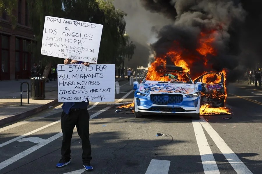 Un activista sostiene carteles mientras un coche arde durante las protestas desatadas por las redadas de inmigración en Los Ángeles, California, Estados Unidos. EFE/EPA/ALLISON DINNER
