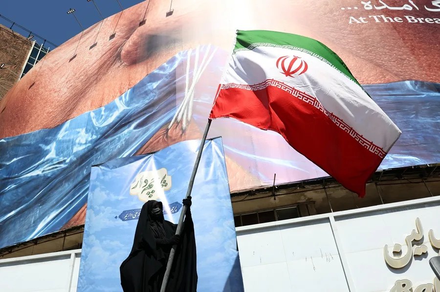 Una mujer velada sostiene la bandera nacional de Irán frente a una enorme valla publicitaria