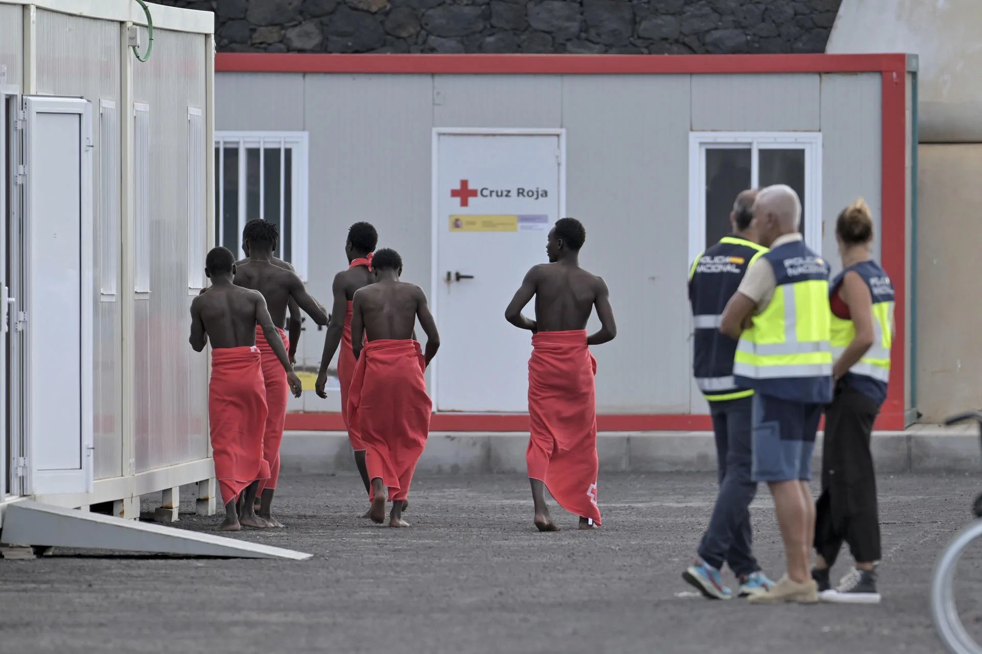 Tres policías observan en el muelle de La Restinga (El Hierro) a cinco de las 119 personas que iban a bordo del cayuco que llegó a la isla el pasado 23 de junio. EFE/Gelmert Finol