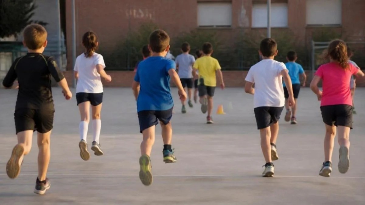 Grupo de ni&ntilde;os y ni&ntilde;as corriendo en una pista escolar, vistos de espaldas, hacia unos conos