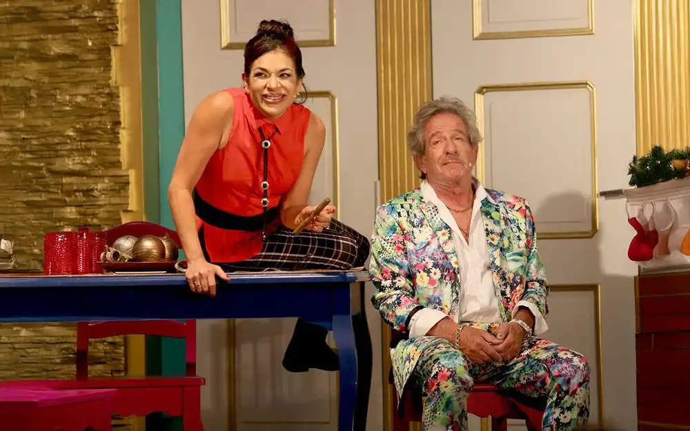 Claribel Medina, sobre la mesa, y Pablo Alarcón, sentado con gesto serio, durante una escena de su obra "Es complicado".