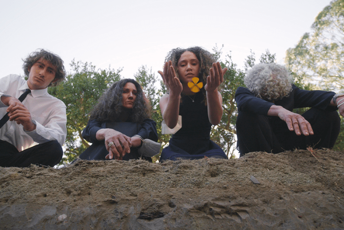 Four people viewed from below standing on a dirt surface, with trees in the background, one releasing a yellow flower.