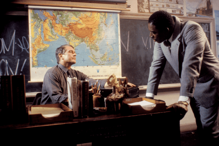 Two men engage in a serious discussion in a classroom, with one seated at a desk and the other standing beside him in a light gray suit. A map of Asia is prominently displayed behind them.