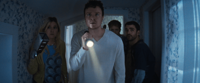 Four characters in a dimly lit hallway, with one man holding a flashlight, illuminating his concerned expression.