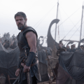 A muscular warrior in dark armor stands on a beach, gazing intently, with a fleet of ships and a camp visible in the background.