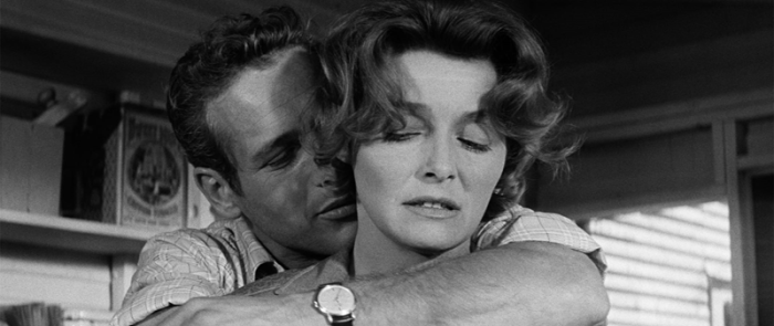 A black-and-white still from "HUD," showing a man embracing a woman in a domestic setting; the woman appears pensive with her eyes closed while the man leans in closely.