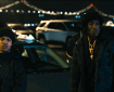 Two individuals in black clothing stand beside a parked silver car at night, with a lit bridge and city lights in the background.