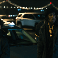 Two individuals in black clothing stand beside a parked silver car at night, with a lit bridge and city lights in the background.