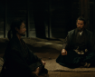 A dimly lit scene featuring two men seated on a mat in a prison cell, one in a dark robe meditating and the other in a samurai outfit, appearing contemplative.