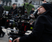 A crew member operates a professional camera on set, wearing a black beanie and coat, with blurred crew members in the background.