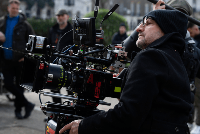A crew member operates a professional camera on set, wearing a black beanie and coat, with blurred crew members in the background.