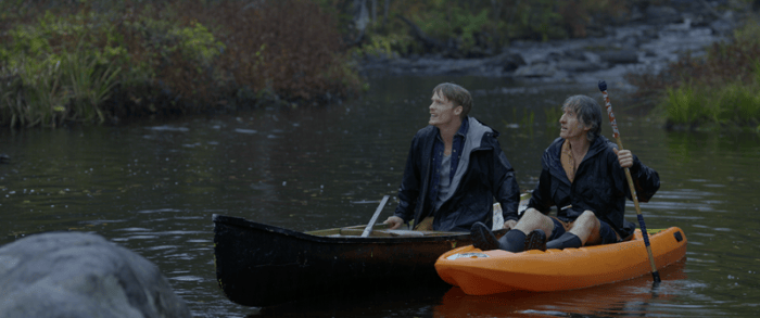 Two men in a black canoe and an orange kayak float on a tranquil river, surrounded by greenery, engaging in quiet observation.