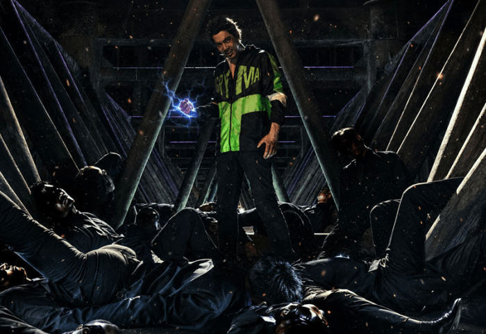 A man in a bright green and black jacket stands confidently over figures in black clothing, gripping a glowing sphere of energy in a dark industrial setting.