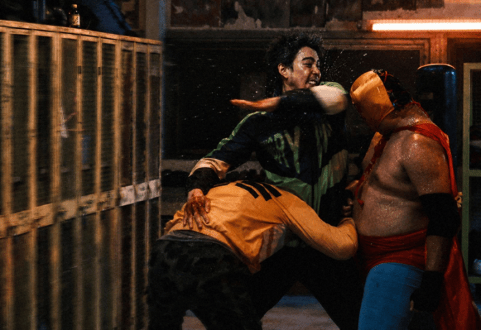 A wrestling scene from "GOD SKIN" showing three men in a vigorous confrontation, one striking another while a third man grapples below.