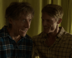 Two male characters sit closely together, engaged in conversation, with warm yellow curtains in the background.