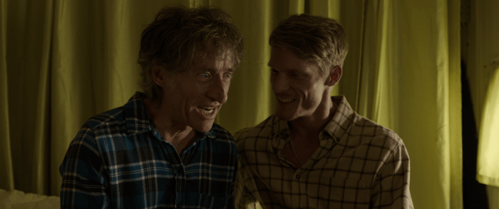 Two male characters sit closely together, engaged in conversation, with warm yellow curtains in the background.