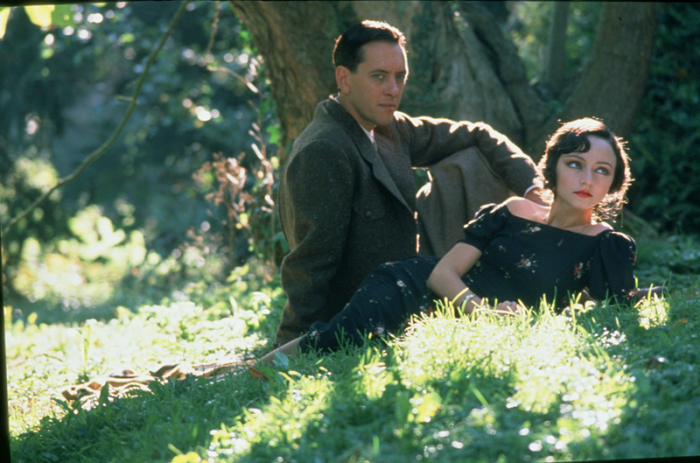 A woman in a dark floral dress lies in green grass, while a man in a tailored coat sits against a tree, both illuminated by dappled sunlight.