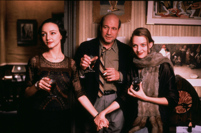 Three people at a gathering: a woman in a black dress holding a glass, a man in a jacket with a glass, and a woman in a fur scarf holding a jar, all standing close together.