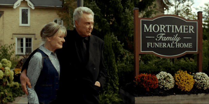 A man and woman stand together in front of a "Mortimer Family Funeral Home" sign, surrounded by greenery and flowers.