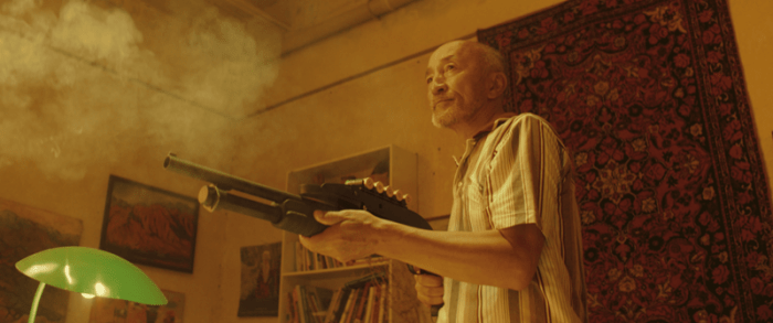 An older man in a striped shirt stands in a dimly lit room holding a weapon, surrounded by smoke and warm lighting.