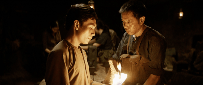 A young man holds a torch while an older man observes him in a dark, underground setting from the film TUNNELS: SUN IN THE DARK.
