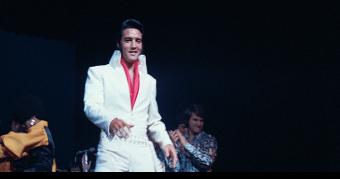 Elvis Presley performing in a white jumpsuit with a red shirt, surrounded by other musicians on stage in a dimly lit concert.