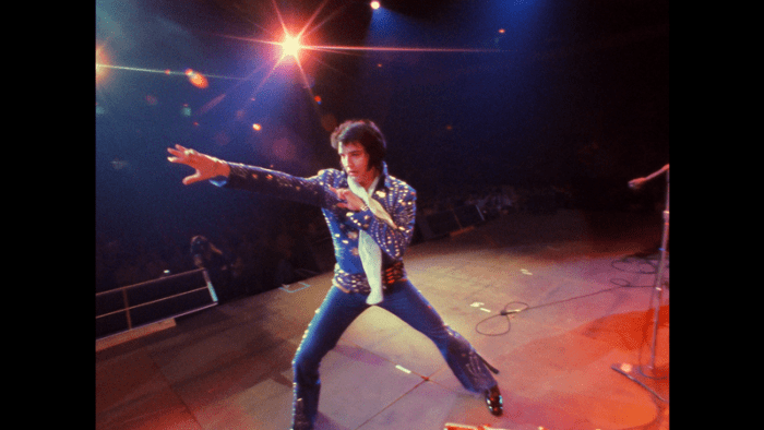Elvis Presley performs on stage in a blue sequined jumpsuit, striking a dynamic pose with colorful stage lights in the background.