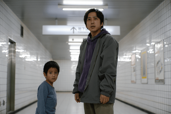 A young boy and a man stand in a brightly lit subway station, both looking concerned towards the camera. The boy wears a light blue shirt, while the man is dressed in a gray jacket over a purple sweatshirt.
