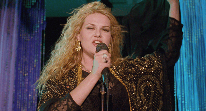 A woman with long blonde hair performs on stage, wearing a black sequined outfit and large star-shaped earrings, holding a microphone and raising her arm.
