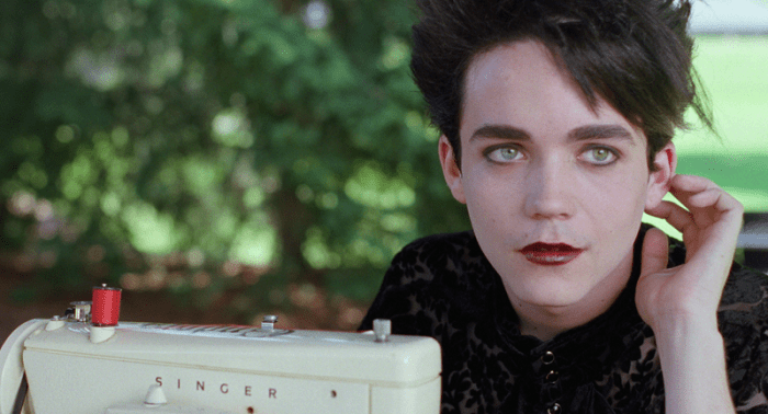 A young person with dark makeup and a burgundy lip color sits next to a vintage Singer sewing machine in a green outdoor setting, their hand touching their neck.