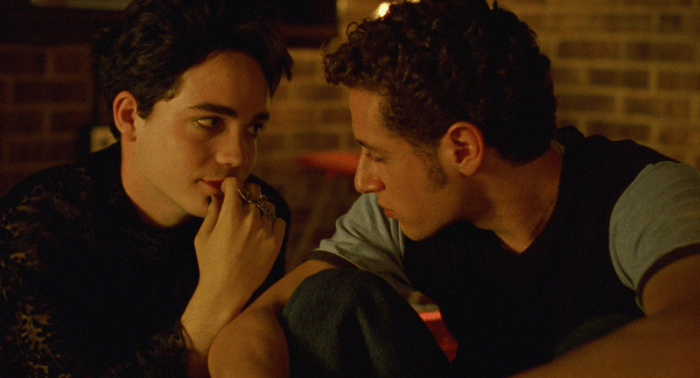 Two young men sit closely together in a dimly lit room, engaged in a thoughtful conversation, with one holding their finger to their lips.