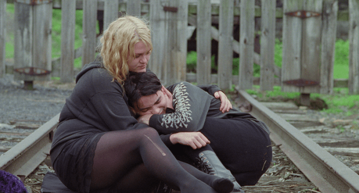 Two individuals sitting on railway tracks, one with blonde hair comforting another with black hair, both in dark clothing, in a green outdoor setting.