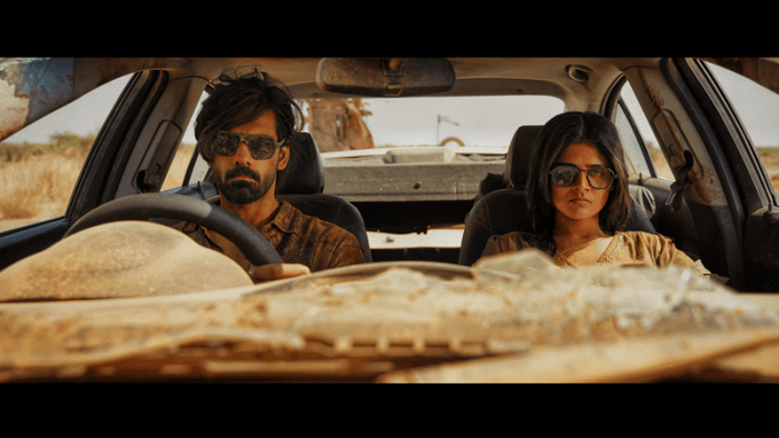 A man and woman sit in a dusty car, both wearing sunglasses. The man, in a brown shirt, drives with a serious expression, while the woman, in a light top, looks contemplative. The exterior shows an arid landscape.