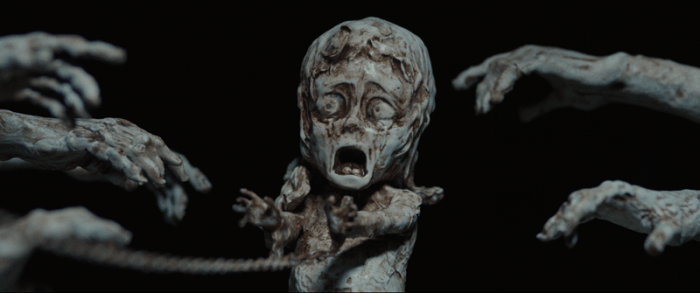 A frightened, pale figure with a wide-open mouth is surrounded by skeletal hands reaching out toward it, set against a black background.