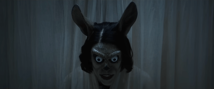 A character with a rabbit-like mask, featuring large eyes and ears, peering out from behind sheer curtains.