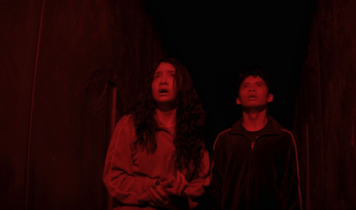 A still from the film HOME BODIES showing a concerned woman and a surprised man in a narrow, red-lit corridor.