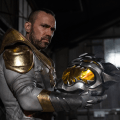 A man in the White Dragon costume holds a metallic helmet resembling a dragon's head in a dimly lit industrial setting.