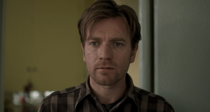 Ewan McGregor in Beginners, close-up with a thoughtful expression, wearing a plaid shirt, set in a blurred domestic background.