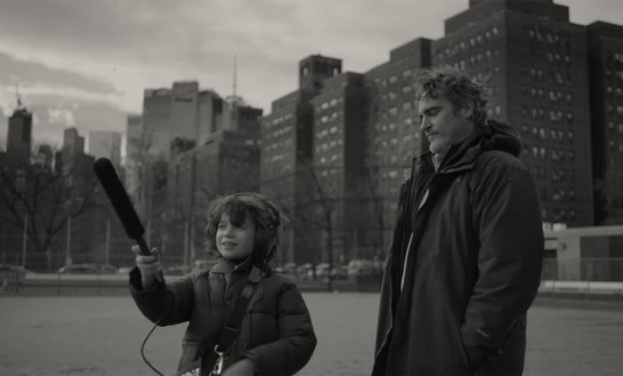 A young boy holds a microphone on a boom pole next to an adult man in an urban setting, both dressed in dark coats, with tall buildings and an overcast sky in the background.
