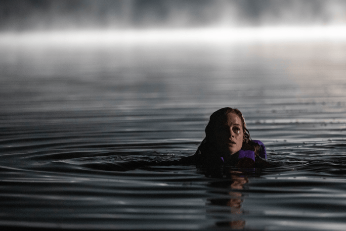 A young woman in a purple life jacket floats in calm water at night, her expression contemplative, surrounded by a misty backdrop.