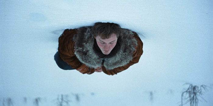 A person lies on snow-covered ground, wearing a brown fur-lined jacket, viewed from an inverted perspective.
