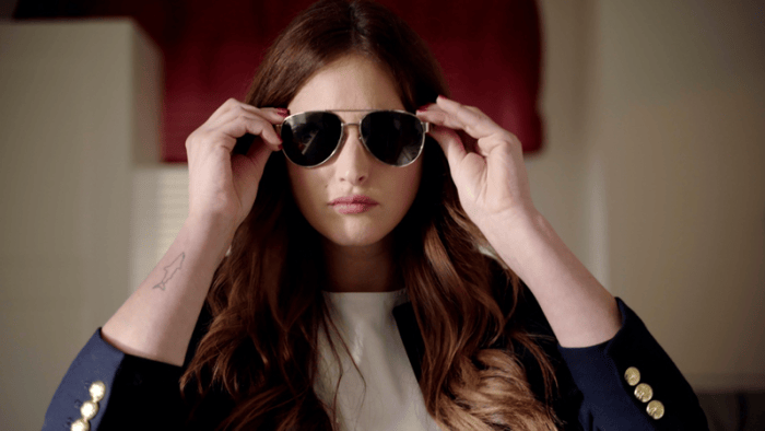 Woman in a dark blazer adjusting aviator sunglasses indoors.