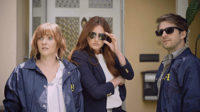 Three adults stand outside a building, with two wearing sunglasses and all looking toward something off-camera.