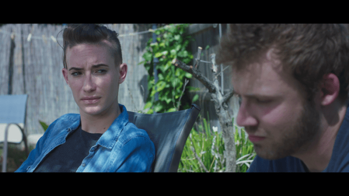Film still of a woman in a denim jacket looking seriously at a bearded man seated beside her outdoors.