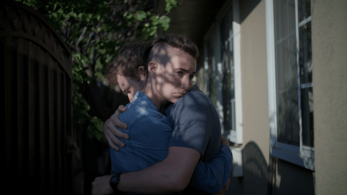 Two people embrace in a sun-dappled passage beside a house.